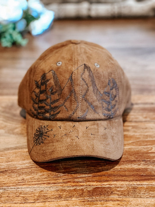 Baseball hat with hand-drawn mountains, fir trees, and a dandelion on the bill shown in brown