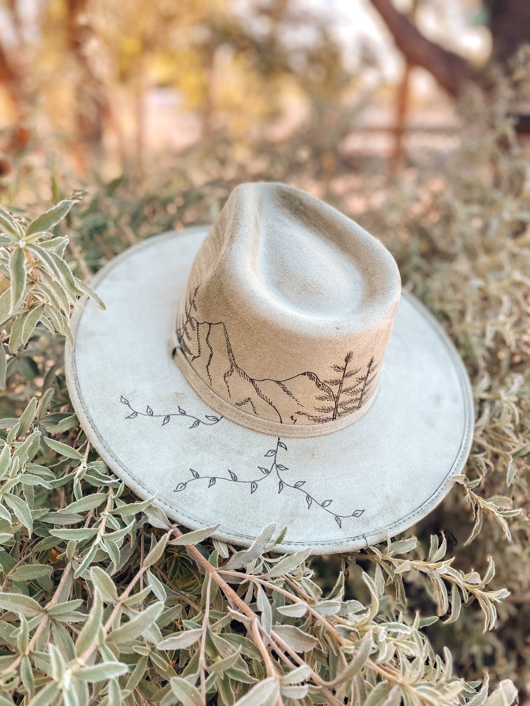 Neutral fedora with hand-drawn mountain, fir tree, and vine design shown in sage green