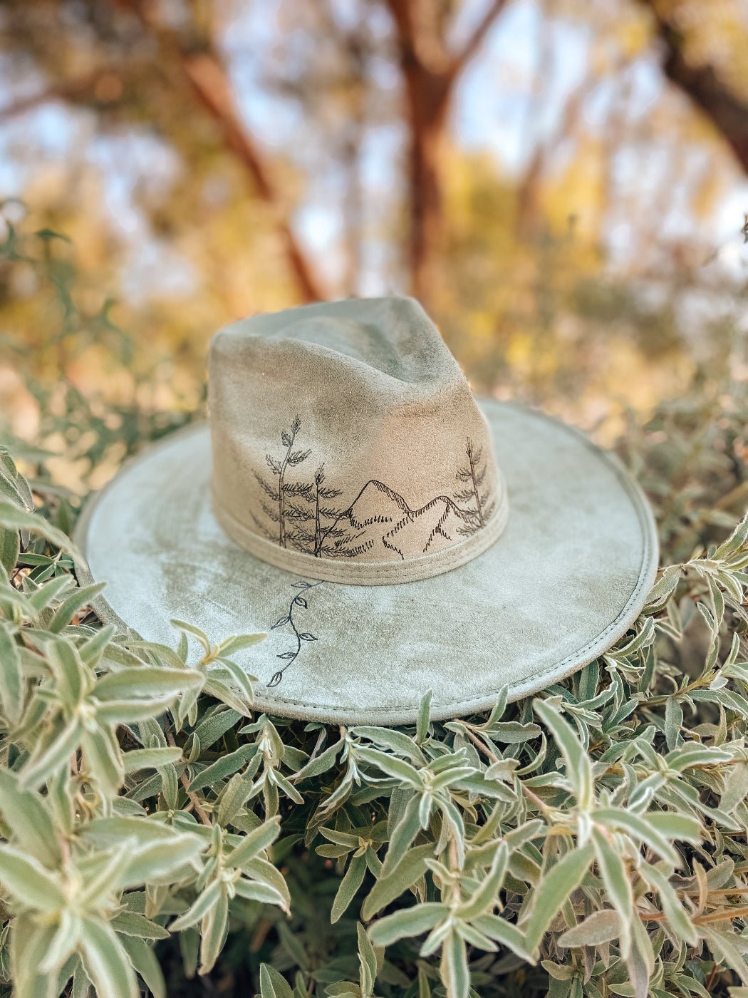 Neutral fedora with hand-drawn mountain, fir tree, and vine design shown in sage green